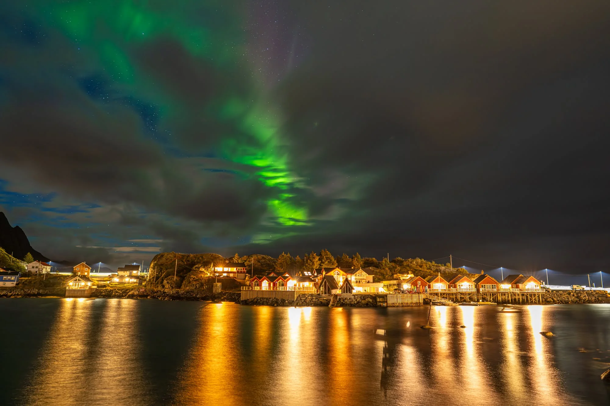 Northern lights over the lit-up red cabins of Hamnøy, Lofoten, reflected in the water