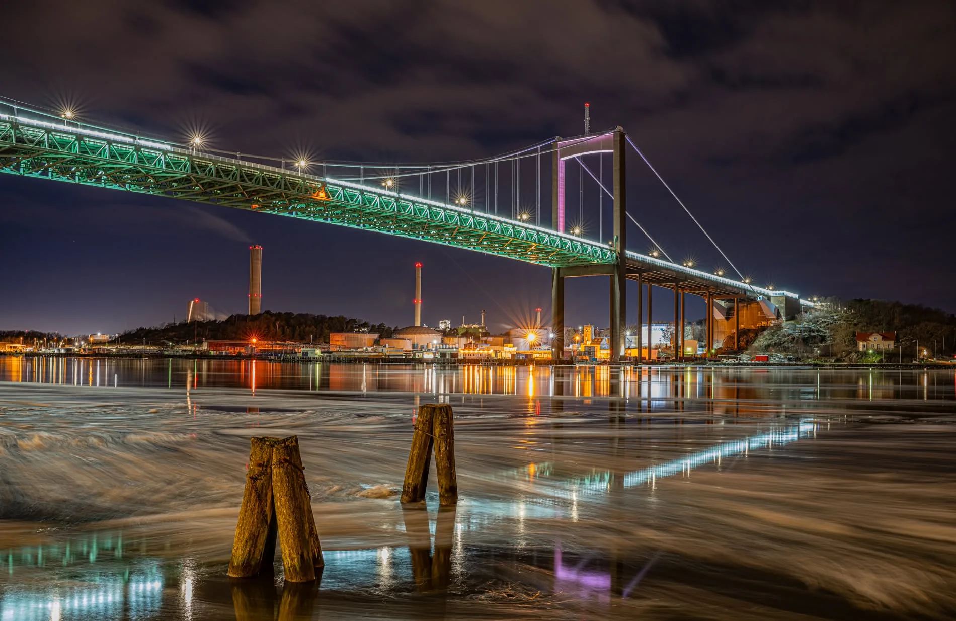 Ice floes drifting past old wooden mooring posts under the lit Älvsborg Bridge at night, Port of Gothenburg