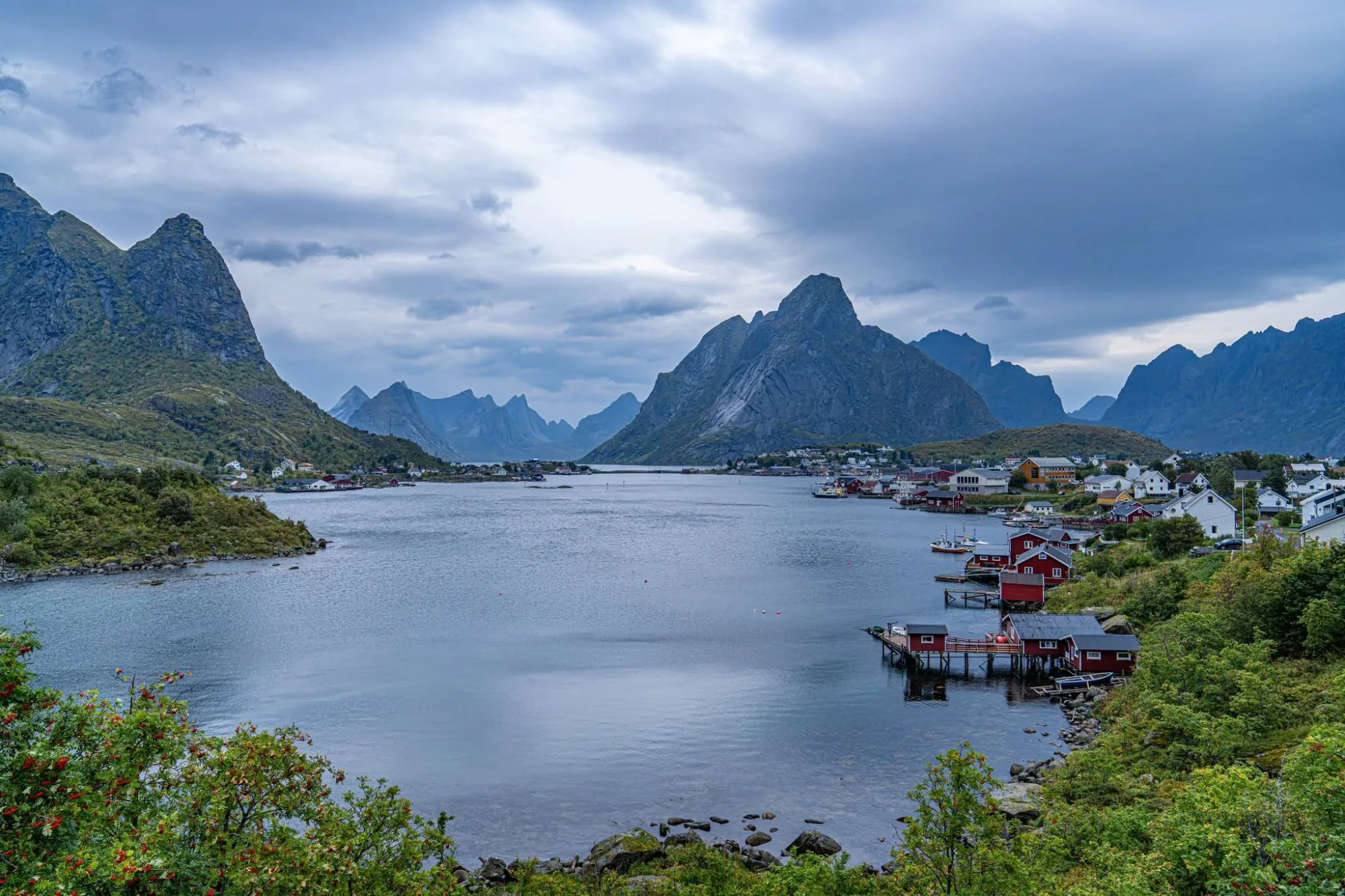 Reine in Lofoten, red fishermen's cabins on stilts by a calm bay below jagged mountain peaks and heavy clouds